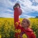 Woman in a red Cavemantraining hoodie holding a kettlebell overhead in a yellow canola field