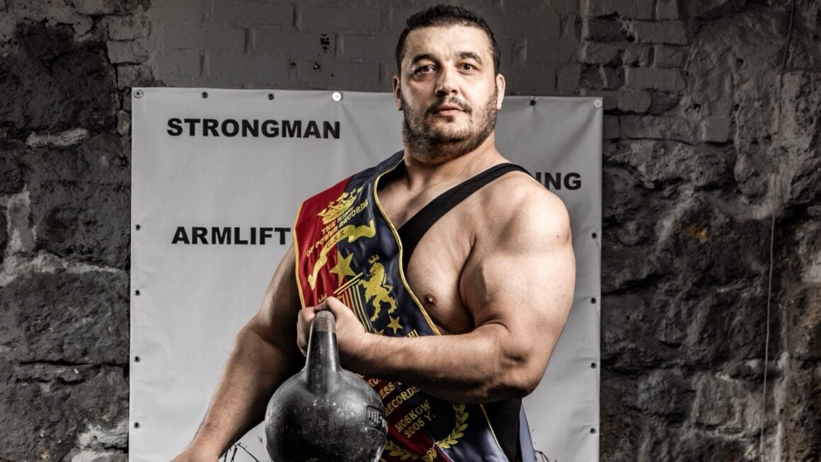 Strongman in a competition sash holding an old cast-iron kettlebell in a stone-walled gym, beside a loaded barbell