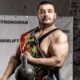 Strongman in a competition sash holding an old cast-iron kettlebell in a stone-walled gym, beside a loaded barbell