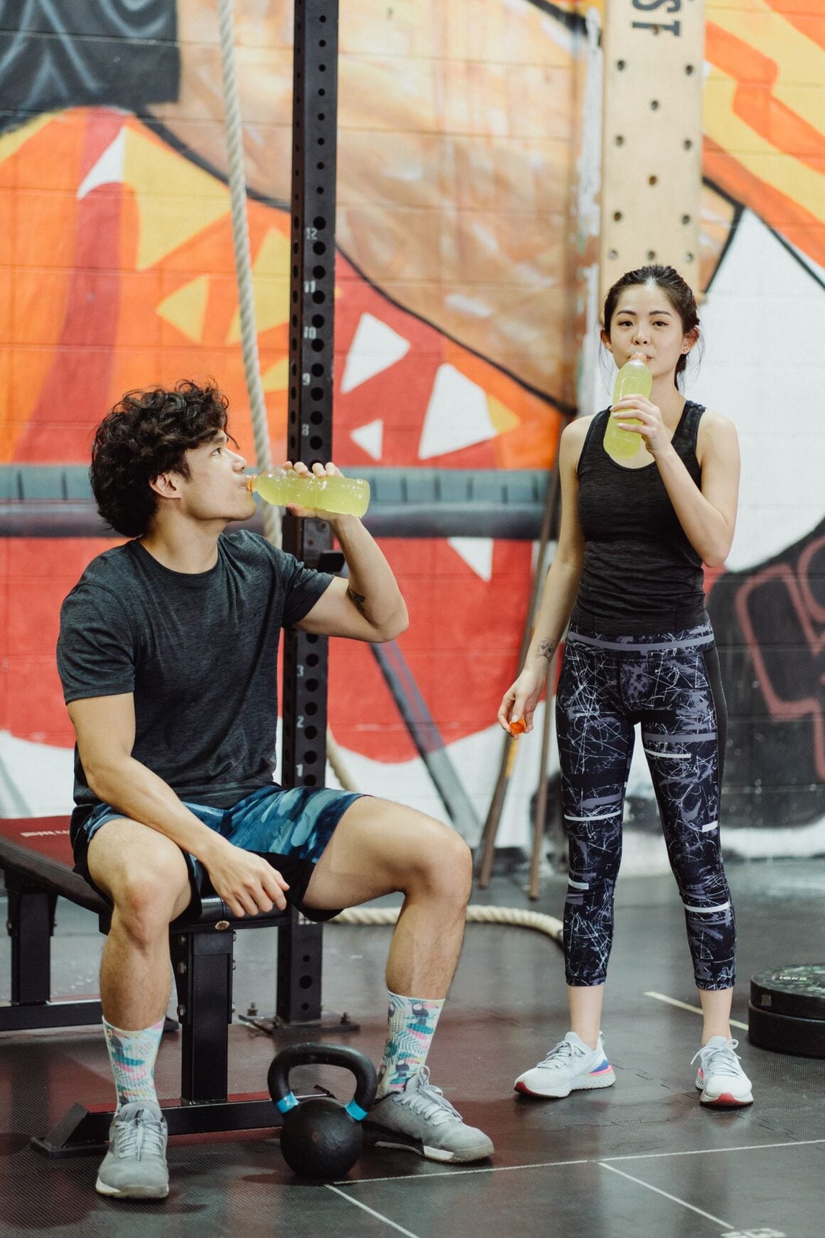 Man sitting on a gym bench drinking from a bottle between sets with a kettlebell at his feet, woman drinking behind him
