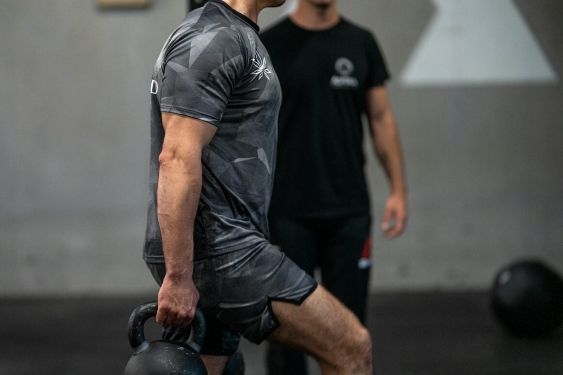 Man holding a kettlebell in a gym while a trainer watches