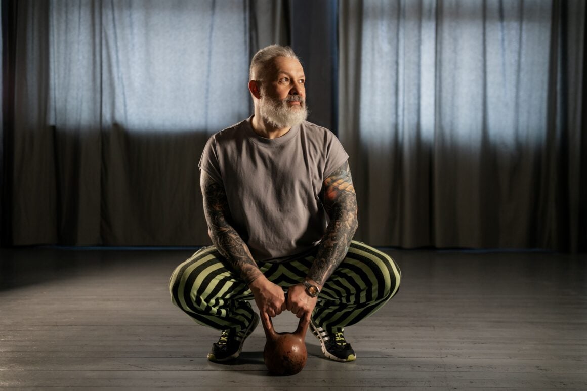 Grey-bearded man in his fifties squatting with a kettlebell between his feet in a dark studio
