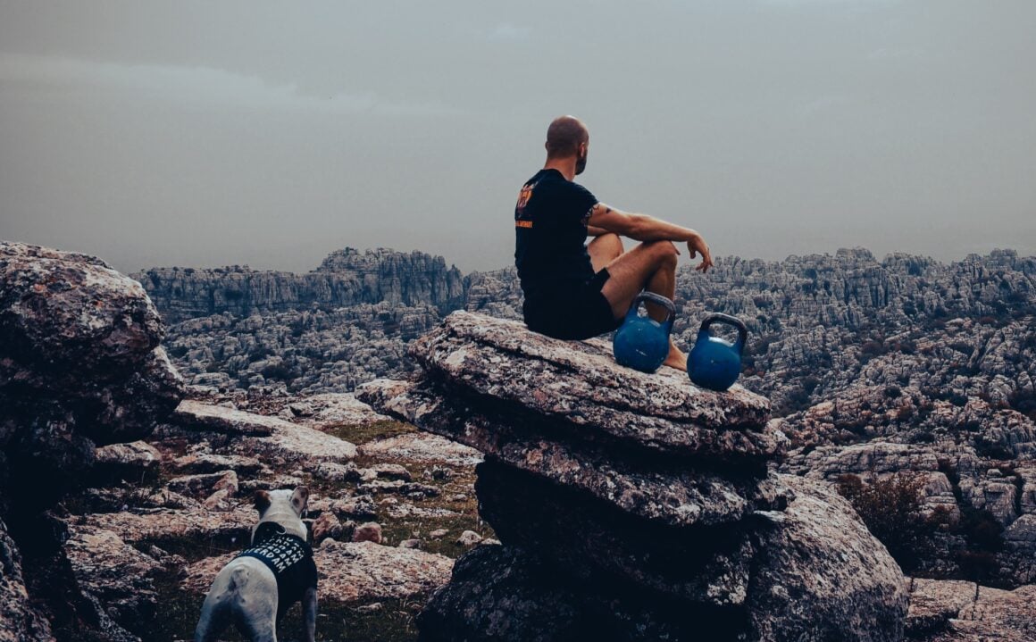 Taco Fleur sitting on a rock ledge at El Torcal with two kettlebells, his dog watching from below