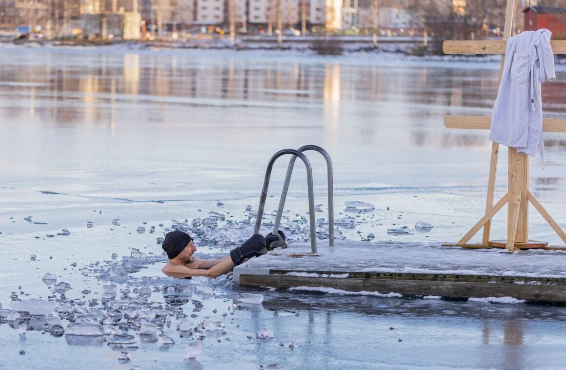 Man in icy lake water holding a dock ladder, with a white bathrobe hanging on a wooden frame beside him