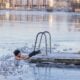 Man in icy lake water holding a dock ladder, with a white bathrobe hanging on a wooden frame beside him