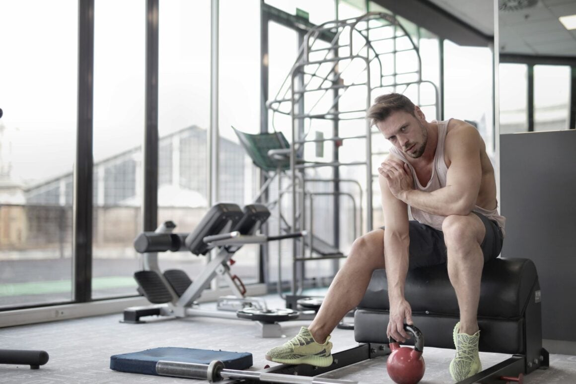 Tired man sitting on a gym bench between sets, head resting on his hand, holding a red kettlebell on the floor