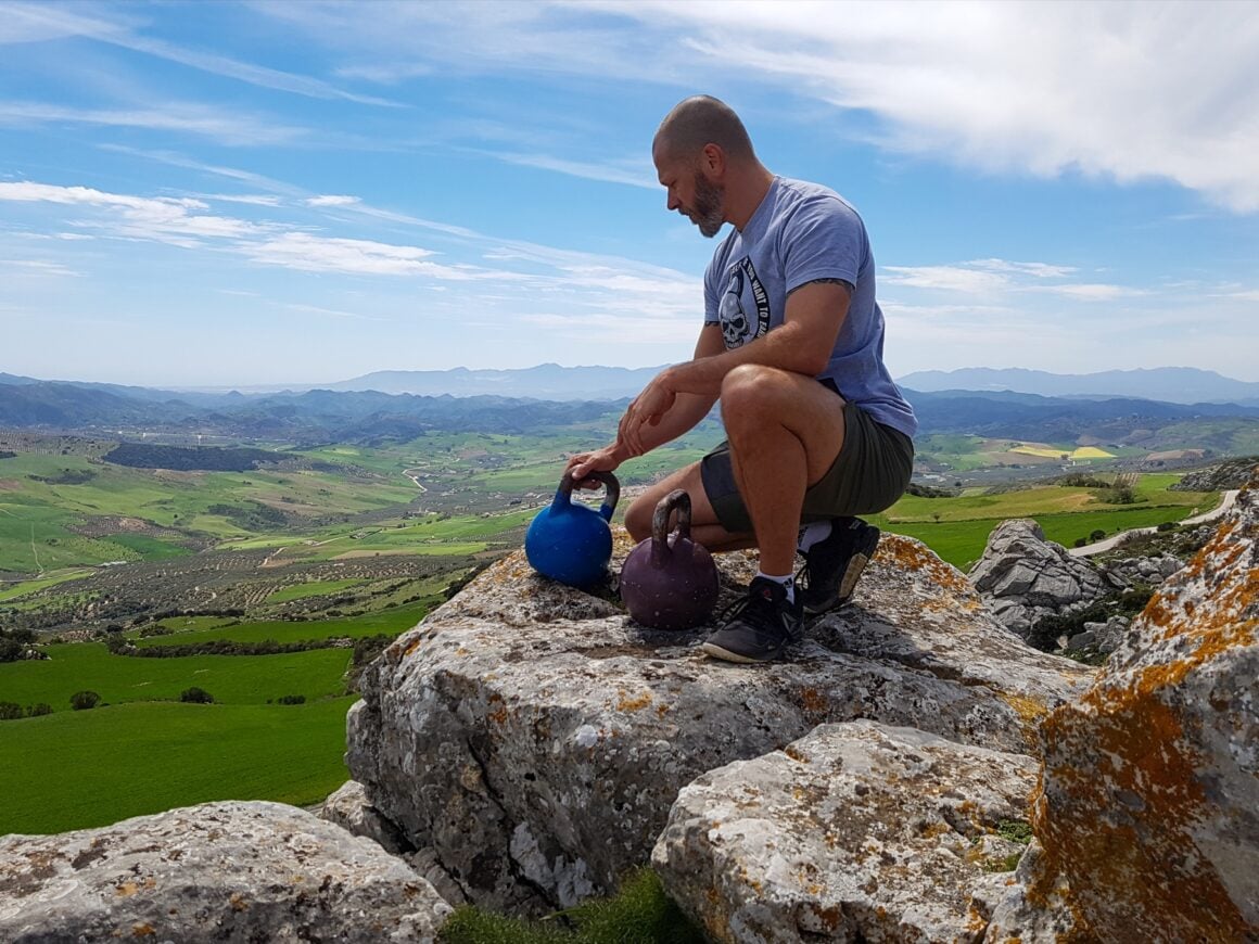 Man sitting beside two kettlebells on a rocky mountain overlook