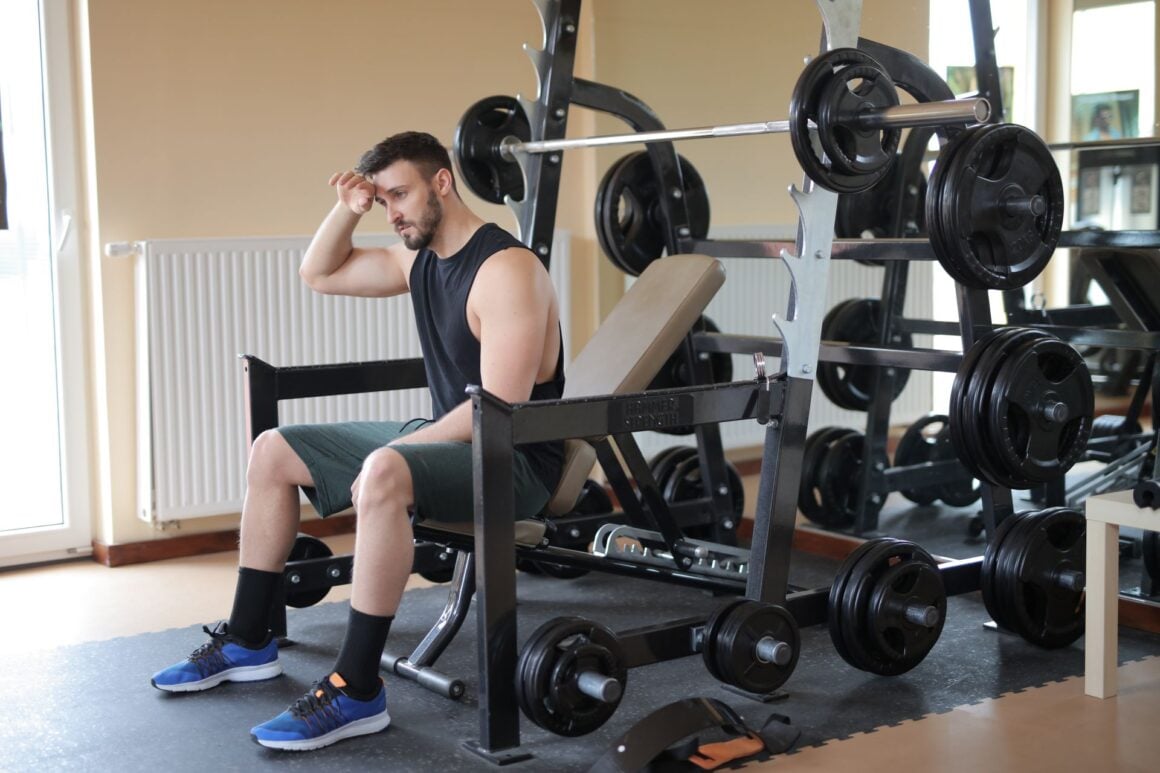 Tired man in a black tank top resting on a weight bench between sets in a home gym