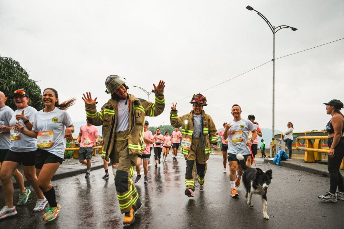 Two firefighters in full turnout gear running a rainy community race with smiling runners and a border collie