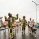 Two firefighters in full turnout gear running a rainy community race with smiling runners and a border collie